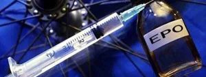 Siringa pronta a prelevare eritropoietina (EPO) da fiala davanti a ruota di bicicletta su sfondo blu, simbolo del doping nel ciclismo.