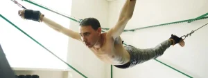 Uomo sospeso in aria in posizione orizzontale tramite funi elastiche, con torso scoperto e sguardo concentrato, in una sala per attività fisiche.