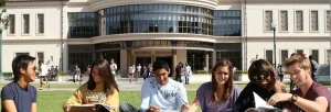 Sei studenti universitari seduti sull’erba nel campus, sorridenti e intenti a studiare con libri e quaderni aperti, davanti all’ingresso di un edificio moderno con vetrate e colonne.