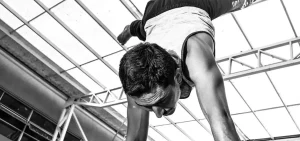 Persona in posa acrobatica a testa in giù, sostenuta sulle mani, con canottiera e pantaloni sportivi. Sfondo con travi metalliche e tetto trasparente. Immagine in bianco e nero.
