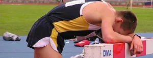 Atleta in abbigliamento sportivo blu e giallo, appoggiato con la testa e le braccia su una barriera di una pista di atletica. Sembra stanco. Sullo sfondo: campo da gioco e borse sparse.