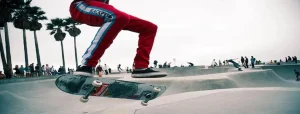Persona che fa skateboard in un parco, indossa pantaloni rossi con striscia bianca e azzurra. Sullo sfondo palme e persone che osservano. Atmosfera di svago all’aperto.