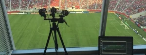 Vista da una cabina di osservazione in uno stadio di calcio con telecamere su treppiede e un computer portatile che monitora la partita.