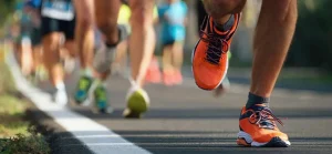 Primo piano di scarpe da corsa arancioni su una strada durante una gara podistica, con altri corridori sfocati sullo sfondo.