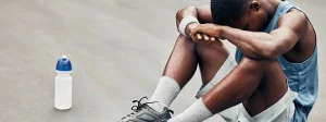 Persona seduta a terra in abbigliamento sportivo, con la testa appoggiata sulle braccia e una bottiglia d’acqua accanto, in un momento di riposo dopo l’allenamento.