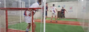 Partita di calcio al coperto con quattro giocatori su campo sintetico, vista dalla porta con rete rossa.