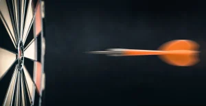 Freccetta arancione in movimento veloce verso il centro di un bersaglio da freccette con sfondo scuro.