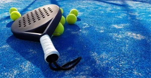 Racchetta da padel nera con manico bianco e palline su campo blu, con ombre ben definite.