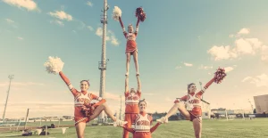 Cinque cheerleader in uniforme rossa e bianca eseguono una coreografia su un campo sportivo con cielo azzurro e nuvole sullo sfondo.