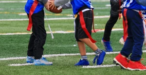 Gruppo di bambini in maglie blu che giocano a flag football su un campo erboso, con bande rosse legate alla vita e palla ovale.