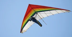 Persona in volo con deltaplano colorato con strisce arcobaleno, cielo blu e limpido sullo sfondo.