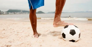 Persona sulla spiaggia con piede appoggiato su un pallone da calcio, mare e montagne sullo sfondo.