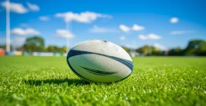 Pallone da rugby posato sull’erba verde di un campo con cielo azzurro e nuvole sullo sfondo.