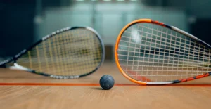 Due racchette da squash poggiate sul pavimento di un campo, con una pallina nera tra di loro e pareti traslucide sullo sfondo.