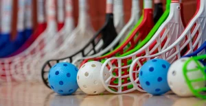 Mazze da floorball di vari colori e palline blu e bianche con fori, disposte su un pavimento.