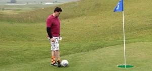 Persona che gioca a footgolf su campo verde, con maglietta rossa e pantaloncini a quadri, pronta a calciare verso una buca con bandierina blu.