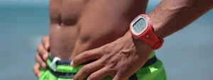 Torso di una persona con orologio sportivo rosso e pantaloncini verdi in un ambiente marino.