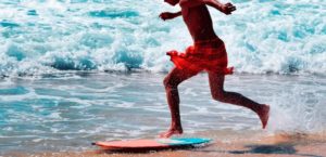 Persona con pantaloncini rossi corre sulla sabbia umida per saltare su una tavola da skimboard vicino all’oceano.