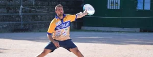 Uomo in maglietta a righe gialle e blu colpisce la palla con un tamburello su un campo all’aperto in terra battuta.