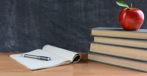 Quaderno aperto con penna su una scrivania in aula scolastica, accanto a tre libri impilati con una mela rossa sopra, e una lavagna sullo sfondo.
