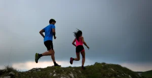 Due persone che corrono su un sentiero di montagna erboso e roccioso, con abbigliamento sportivo: maglietta azzurra e rosa, pantaloncini neri, cielo nuvoloso.