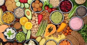 Varietà di alimenti vegetali con ciotole di legumi, cereali, semi, hummus, guacamole, frutta e verdura fresca, pane integrale e tofu.