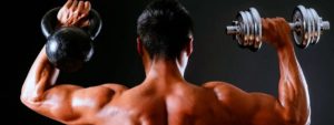 Persona vista di spalle solleva kettlebell e manubrio, con muscoli di schiena e braccia definiti su sfondo scuro.