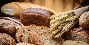 Varietà di pane rustico su un tavolo, con diversi tipi di pane interi e affettati di varie texture e colori, accompagnati da un fascio di spighe di grano.