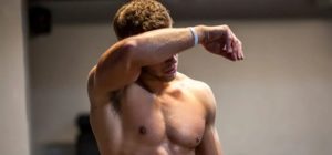 Uomo a torso nudo che si asciuga il sudore dalla fronte con un braccio, in un ambiente interno simile a una palestra