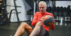 Uomo in palestra con maglia rossa e pantaloncini neri seduto a terra mentre tiene una palla medica, circondato da attrezzi e pesi da allenamento