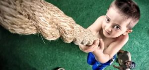 Bambino a torso nudo che si arrampica su una corda spessa sopra un pavimento verde simile a erba sintetica, mentre una persona lo osserva in basso a destra.