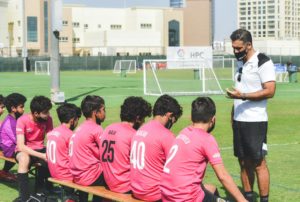 Gruppo di giovani calciatori con maglie rosa seduti su una panchina mentre un allenatore con maschera nera e maglietta bianca dà istruzioni sul campo da calcio.