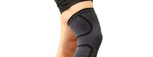 Persona con ginocchiera elastica scura a compressione e pantaloncini neri su sfondo bianco.