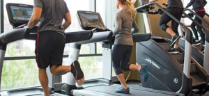 Persone in palestra che si allenano su tapis roulant e macchine simili a scale, con abbigliamento sportivo e luce naturale che entra dalle ampie finestre.