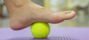 Piede posato su palla da tennis gialla su tappetino viola, esercizio per massaggio e rilassamento muscolare.