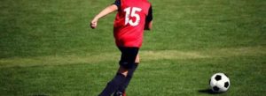 Giocatore con maglia rossa numero 15 e pantaloncini neri in movimento su campo da calcio verde verso un pallone.