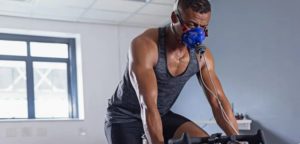 Uomo con canottiera sportiva che pedala su bici stazionaria indossando una maschera respiratoria collegata a sensori, in una stanza luminosa con finestra.