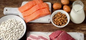 Varietà di alimenti proteici disposti su un tavolo in legno: salmone, uova, latte, mandorle, fagioli bianchi, carne rossa e carne di maiale su taglieri bianchi.
