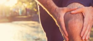 Persona che si tiene il ginocchio con entrambe le mani, segnalando dolore o affaticamento, con sfondo sfocato e riflessi di luce solare.