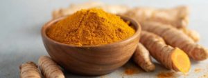 Ciotola di legno con polvere arancione di curcuma e rizomi freschi di curcuma su sfondo chiaro sfocato.