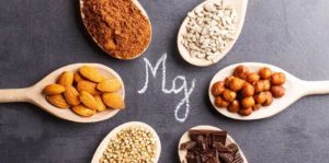 Sei cucchiai di legno disposti a cerchio su sfondo scuro, contenenti alimenti ricchi di magnesio: semi di girasole, nocciole, cioccolato fondente, grano saraceno, mandorle e cacao in polvere. Al centro, la sigla “Mg” scritta con gesso.