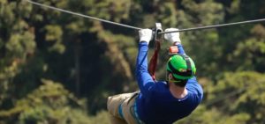 Persona che fa zip-lining nella foresta con casco verde, maglietta blu, guanti bianchi e imbracatura di sicurezza, circondata da vegetazione.