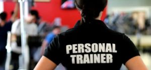 Persona di schiena con maglietta nera “PERSONAL TRAINER” in palestra, con altre persone che si allenano sullo sfondo.