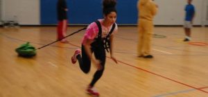 Persona in palestra con maglietta rosa e pantaloni neri, indossa un apparecchio di resistenza; cerchi colorati sul pavimento e altre persone sullo sfondo.