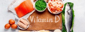 Alimenti ricchi di vitamina D come salmone, pesce intero, uova, formaggio e gamberetti disposti intorno a una tavola di legno con la scritta "Vitamin D".