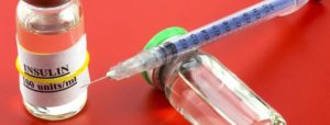Fiala di insulina con etichetta "INSULIN 100 units/ml" accanto a una siringa dosatrice, su sfondo rosso.