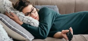 Persona con occhiali e maglione verde scuro sdraiata su un divano, tiene un telecomando e si appoggia a un cuscino soffice; sul divano cuscini di diverse texture e colori.