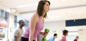 Donna con canottiera viola che solleva un manubrio verde durante una lezione di allenamento con i pesi in palestra, con altre persone sullo sfondo.