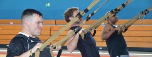 Tre persone si allenano con cinghie di sospensione TRX in palestra, davanti a una parete blu con panche di legno sullo sfondo.