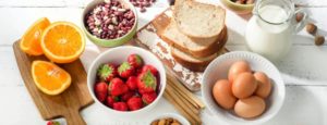 Alimenti su tavolo bianco: pane, latte, uova, fragole, frutta secca, arance tagliate e grissini, disposti in modo ordinato.